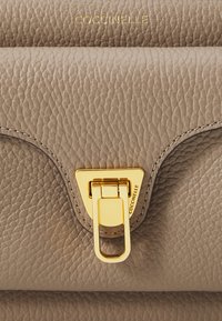 Borsa a tracolla in pelle beige con texture, dotata di una chiusura dorata e logo impressa. Presenta un design curvato e cuciture sottili lungo i bordi.