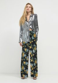 Metallisch silbernes Langarmhemd mit Knopfdesign, kombiniert mit weiten floral gemusterten Hosen aus dunklem Stoff, die gelbe und blaue Blumen zeigen.