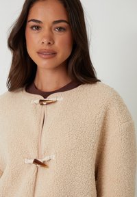 Beige fleece bunda s jemnou texturou, kulatým límcem a uzávěrem na knoflík s kontrastním hnědým akcentem.