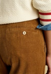 Brūnas kordurojas bikses ar aizmugures kabatu, kurai ir koka pogu, un vertikālu rievotu tekstūru. Džemperis ir krēmkrāsā ar sarkanām svītrām.