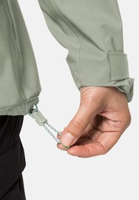 Groene lichtgewicht jas van textuurstof. Beschikt over een verstelbaar trekkoord aan de zoom, bevestigd met een plastic stop. Hand zichtbaar die het koord vasthoudt.