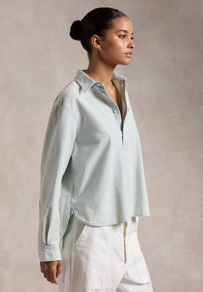 Bunba és világoskék gombos ingben, fehér nadrágban lévő nő, jobbra nézve, semleges háttér előtt.