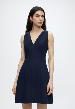 Žena s dlhými tmavými vlasmi, oblečená v tmavomodré bez rukávov s výstrihom do V, stojaca na jednoduchom bielom pozadí, pozriac sa mierne doprava.