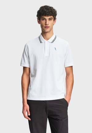 Uomo che indossa una polo bianca a maniche corte con doppie strisce nere sul colletto e un piccolo logo di uccello blu, abbinata a pantaloni neri.