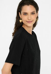 Schwarzes T-Shirt mit einem strukturierten Design und vertikalen Reihen von Verzierungen. Das Modell trägt große goldene Herzohrringe und zeigt einen lässigen Stil.