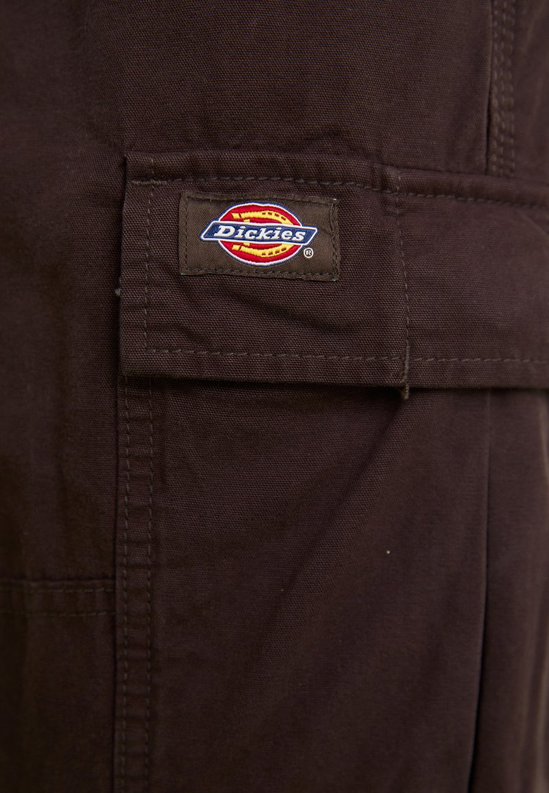 Tissu marron Dickies avec une surface texturée. Présente un patch logo proéminent en rouge, jaune et bleu. Les détails de couture sont visibles.