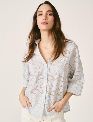 Mujer con cabello largo y castaño, vistiendo una camisa de botones con estampado en azul claro y beige, y pantalones blancos, de pie con las manos en los bolsillos.