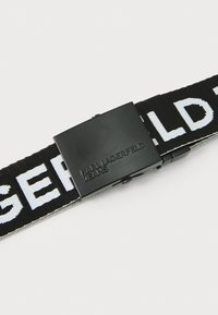 Svart stoffbelte med hvit skrift, med en rektangulær svart metallspenne preget med "KARL LAGERFELD JEANS." Teksturert overflate.