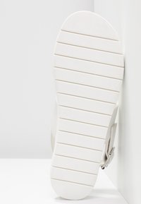 Bílé tenisky s texturovanou podrážkou s horizontálními drážkami, aerodynamickým tvarem a minimalistickým designem. Viditelný detail nastavitelného pásku.