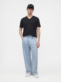 Νέος άνδρας με μαύρο καπέλο, μαύρο T-shirt με V λαιμόκοψη, ανοιχτό μπλε φαρδιά τζιν και λευκά αθλητικά παπούτσια, που στέκεται με το ένα χέρι στην τσέπη.