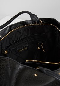 Czarna skórzana torebka z teksturowanym wykończeniem, złotymi akcesoriami oraz czarnym wnętrzem z kieszenią na zamek i detalem z logo.