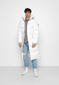 Giovane in piedi contro uno sfondo bianco, indossa un lungo cappotto puffer bianco con cappuccio, jeans strappati blu, camicia a quadri, calzini bianchi e sneakers bianche.