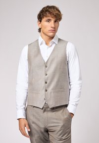 Graue gewebte Weste mit fünf Knöpfen, zwei vorderen Taschen und Revers, getragen über einem weißen Hemd und hellgrauen Hosen.