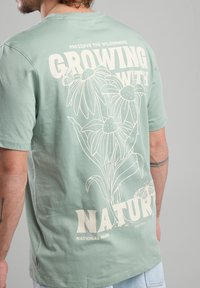 Mintunvihreä puuvillainen t-paita, jossa on valkoinen graafinen kuvasto kukista ja tekstistä "Kasvava luonnon kanssa" takana.