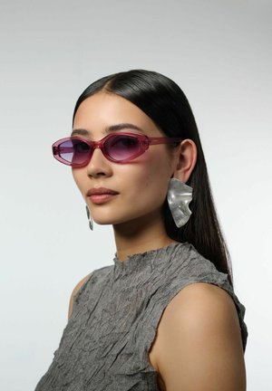 Lunettes de soleil élégantes teintées de violet avec une forme angulaire ; monture transparente. Portées avec un haut gris texturé et de grandes boucles d'oreilles métalliques.