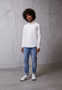 Camicia bianca a maniche lunghe, jeans slim-fit blu chiaro e sneaker bianche. Sfondo in cemento, posa casual, focalizzata sui dettagli dell'abbigliamento.