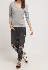 Pull gris à col en V, jean noir déchiré avec bords effilochés, et sandales argentées à design à lanières. Tenue décontractée et minimaliste.