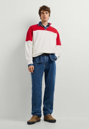 Joven de pie vistiendo un suéter de manga larga rojo y blanco, vaqueros azules y zapatillas marrones sobre un fondo liso y claro.