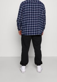 Uomo che indossa una camicia a quadri blu navy, pantaloni jogger neri e sneakers bianche alto, in piedi di fronte a una parete grigia chiaro semplice.