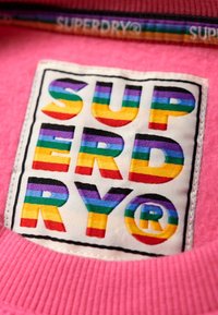 Pink garment label featuring the word "SUPERDRY" in bold, multi-colored letters, with a textured fabric background and black stitching outline.