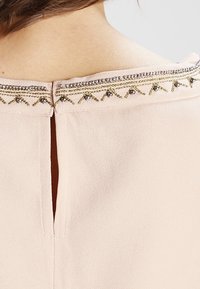 Blusa rosa chiaro con scollo rotondo. Presenta ricami decorativi dorati e argentati lungo il colletto e un'apertura a chiave sul retro.