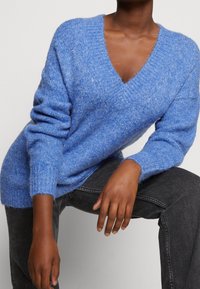 Person som bär en strukturerad blå V-halsad tröja och mörkgrå jeans, sitter med ett knä upphöjt och händerna vilande på benen.