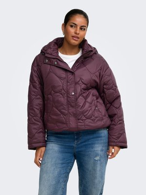 Donna con giacca trapuntata viola con cappuccio e jeans blu, in piedi con le mani lungo i fianchi, guarda leggermente verso destra.