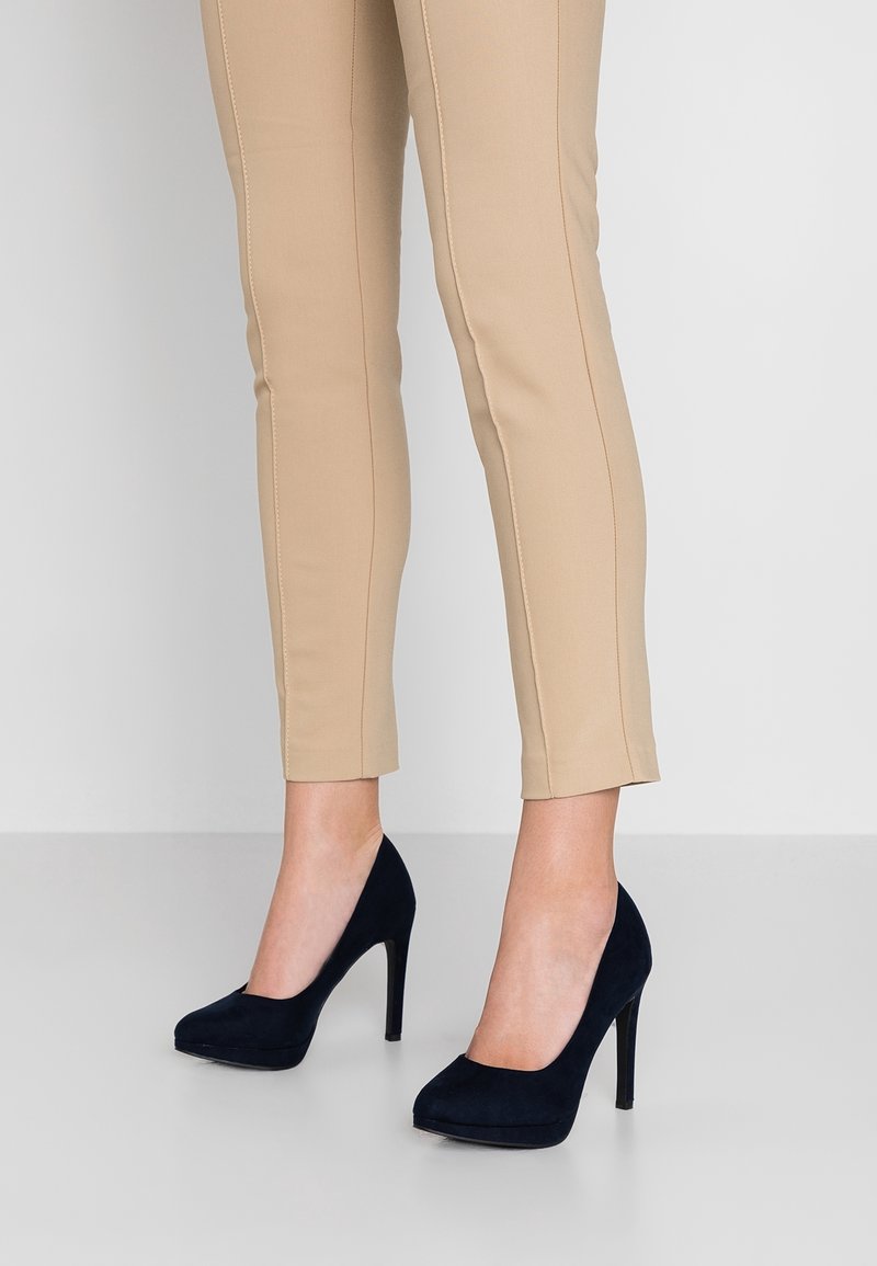 Femme portant un pantalon slim beige et des chaussures à talons noirs, debout sur un sol et un fond clairs et simples.