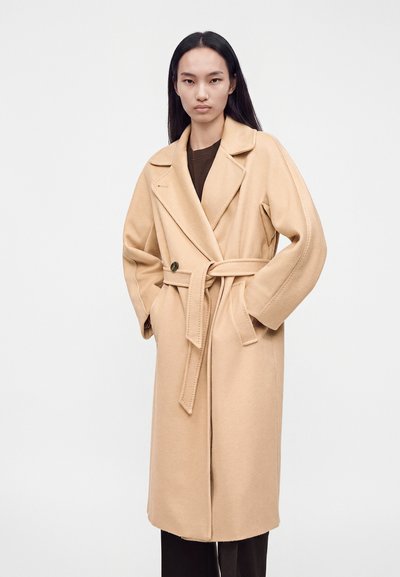 Femme aux longs cheveux foncés portant un manteau en laine beige ceinturé par-dessus un haut sombre, debout, les mains dans les poches du manteau, devant un fond uni.