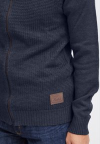Pull en laine bleu marine zippé avec un tissu texturé, des poignets et un ourlet côtelés, et une étiquette en cuir marron portant le nom de la marque "Solid."
