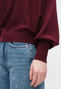 Bordowy sweter z dzianiny o luźnym kroju i opuszczonych ramionach, zestawiony z jasnoniebieskimi podniszczonymi dżinsami z widocznym detalem kieszeni.