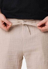 Pantalons beige clair en tissu texturé, dotés d'une taille élastique à cordon, avec un nœud visible et des détails plissés.