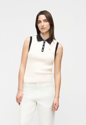 Kobieta ubrana w białą bezrękawnikową dzianinową bluzkę z czarnym kołnierzykiem i guzikami, w połączeniu z białymi spodniami, stojąca na neutralnym tle.