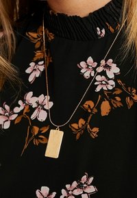 Goldfarbener rechteckiger Anhänger-Halskette auf einer dünnen Kette, getragen über einem schwarzen Oberteil mit floralem Muster in Pink und Braun.