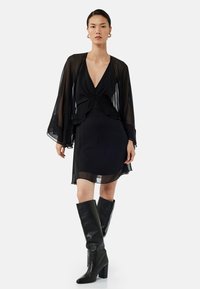 Robe noire avec des manches longues transparentes, décolleté plongeant, design en couches et ourlet au genou. Associée à des bottes noires à talons montantes.