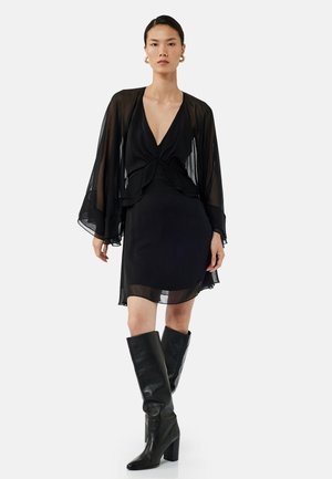 Robe noire avec des manches longues transparentes, décolleté plongeant, design en couches et ourlet au genou. Associée à des bottes noires à talons montantes.