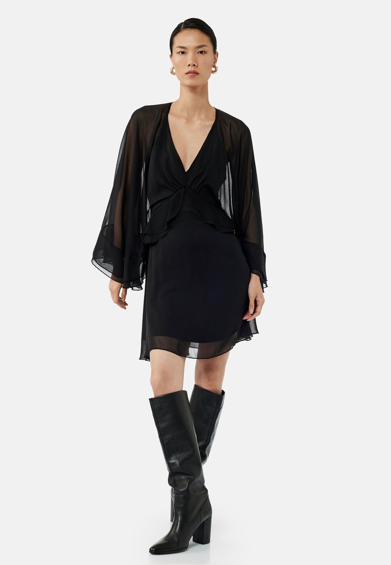 Robe noire avec des manches longues transparentes, décolleté plongeant, design en couches et ourlet au genou. Associée à des bottes noires à talons montantes.