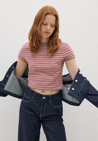 Jonge vrouw met rood haar, gekleed in een rood-wit gestreepte cropped shirt en donkerblauwe jeans, die een blauwe spijkerjack aantrekt.