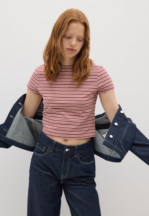 Junge Frau mit rotem Haar, die ein rot-weiß gestreiftes, kurzes Shirt und dunkelblaue Jeans trägt und gerade eine blaue Jeansjacke anzieht.