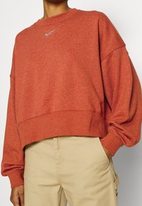 Persona che indossa un felpa Nike di colore ruggine oversize con un piccolo logo e pantaloni beige, con una mano in tasca.