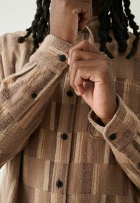 Brunternet skjorte i struktureret stof. Har sorte knapper og en knapmanschet. Mønstret inkluderer varierende nuancer af tan og creme striber.