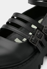 Czarny skórzany but z wieloma wąskimi paskami. Posiada prostokątne klamry oraz grubą, teksturowaną podeszwę zapewniającą przyczepność. Gładkie, błyszczące wykończenie.