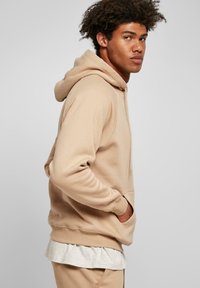 Felpa beige con cappuccio in tessuto morbido, caratterizzata da una tasca frontale, polsini e orlo a costine. Indossata sopra una camicia a strati grigia chiara.