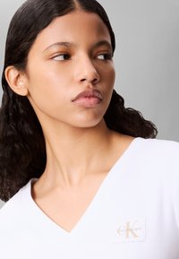 Mujer joven con cabello negro ondulado de longitud media que lleva una camiseta blanca con cuello en V y un pequeño parche con el logo de Calvin Klein.