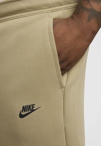 Beige bomullssvettsbyxor med sidficka, med en svart Nike-logotyp på vänster lår och slät yta.