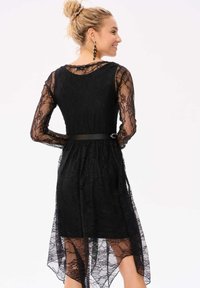 Zwarte kanten jurk met lange, doorzichtige kanten mouwen, een getailleerde lijfje en een uitlopende, hoge-laag zoom. Voorzien van een zwarte riem in de taille.