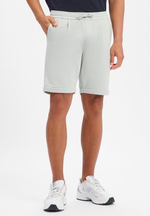 Mężczyzna ubrany w jasnoszare spodenki z trokiem i białe buty sportowe, stojący z jedną ręką w kieszeni na białym tle.