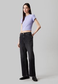 Camiseta de manga corta en color lavanda con corte cropped y detalle bordado, combinada con vaqueros negros de tiro alto y pierna recta, y zapatos negros.