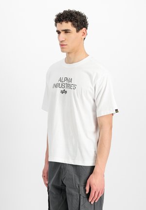 Man with curly hair wearing a white "Alpha Industries" T-shirt and dark cargo pants, standing against a plain white background.