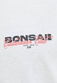Baltas audinys su juodais ir rožiniais tekstais, kuriuose rašoma "Bonsai Cultivation Club" su "Kyoto - Japonija" ir japonų simboliais žemiau. Tekstas turi retro dizainą.